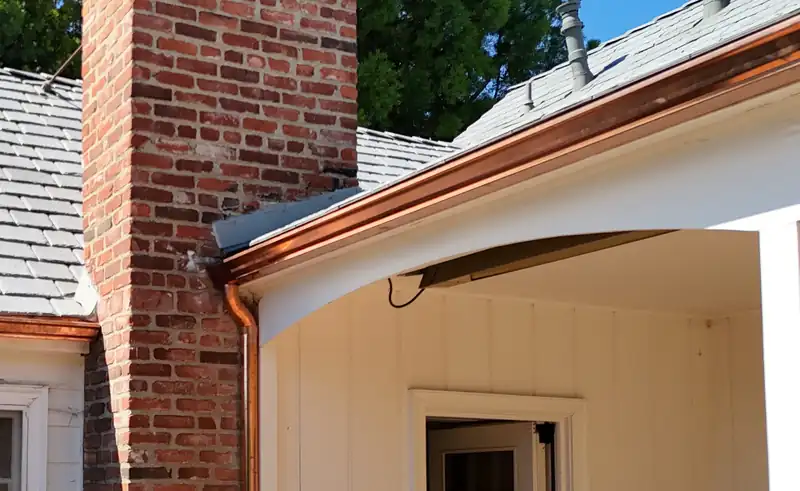 Copper gutter turning the corner of a curved porch eave, with a round copper downspout beside a brick chimney